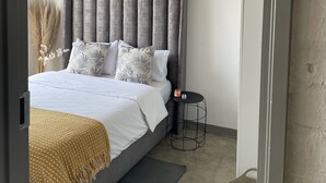 Een slaapkamer met een grijs bekleed hoofdbord, een wit beddeken en een mosterdgele dekentje. Er staat een klein zwart nachtkastje met een kaars en een decoratieve vaas met gedroogde bloemen.