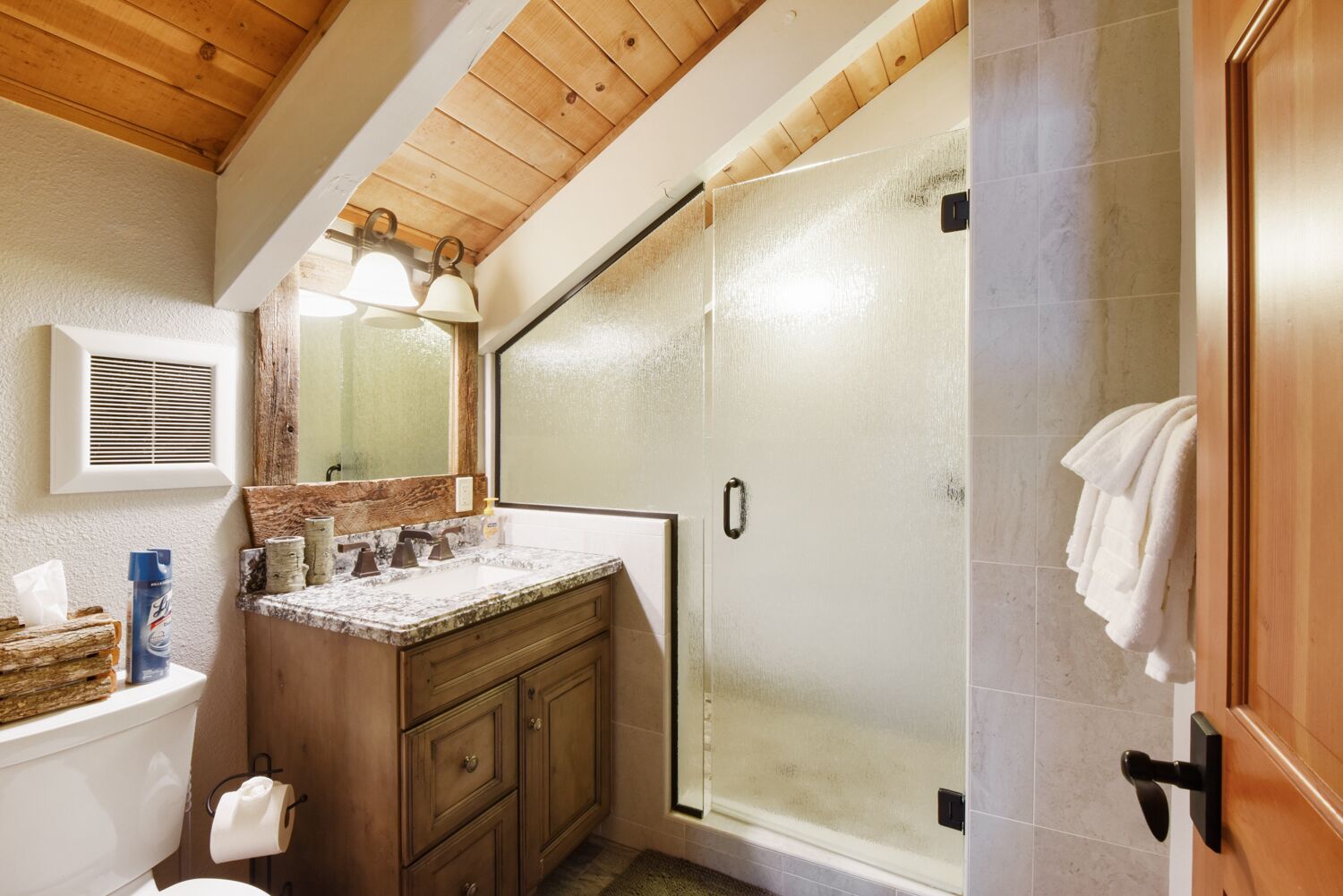 Un baño con techo de madera, cabina de ducha con vidrio, lavamanos de mármol y un inodoro.