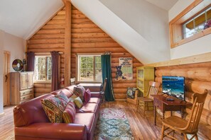A cozy log cabin interior with a leather sofa, wooden furniture, a television, and a scenic view through the windows.