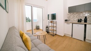 Ett modernt vardagsrum med en grå soffa, ett litet runt bord och en köksdel med tv, skåp och en kaffemaskin.