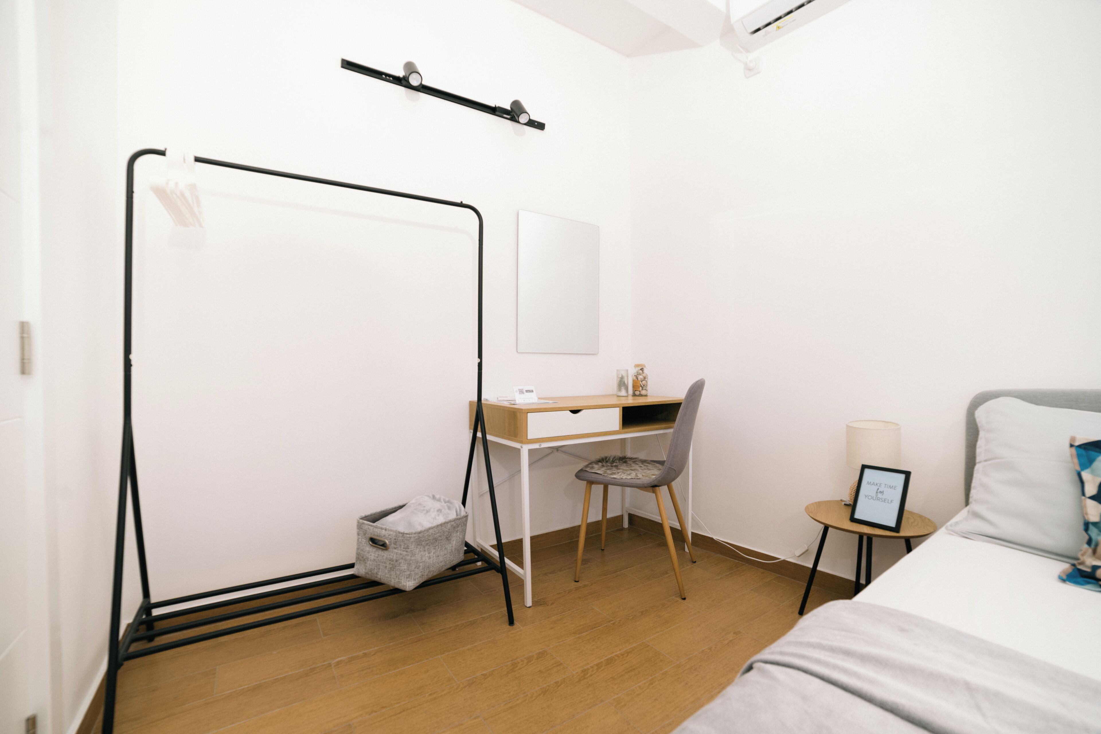 A bedroom with a bed, a desk, a chair, a bedside table, a lamp, and a wall-mounted shelf.