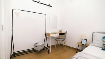A bedroom with a bed, a desk, a chair, a bedside table, a lamp, and a wall-mounted shelf.