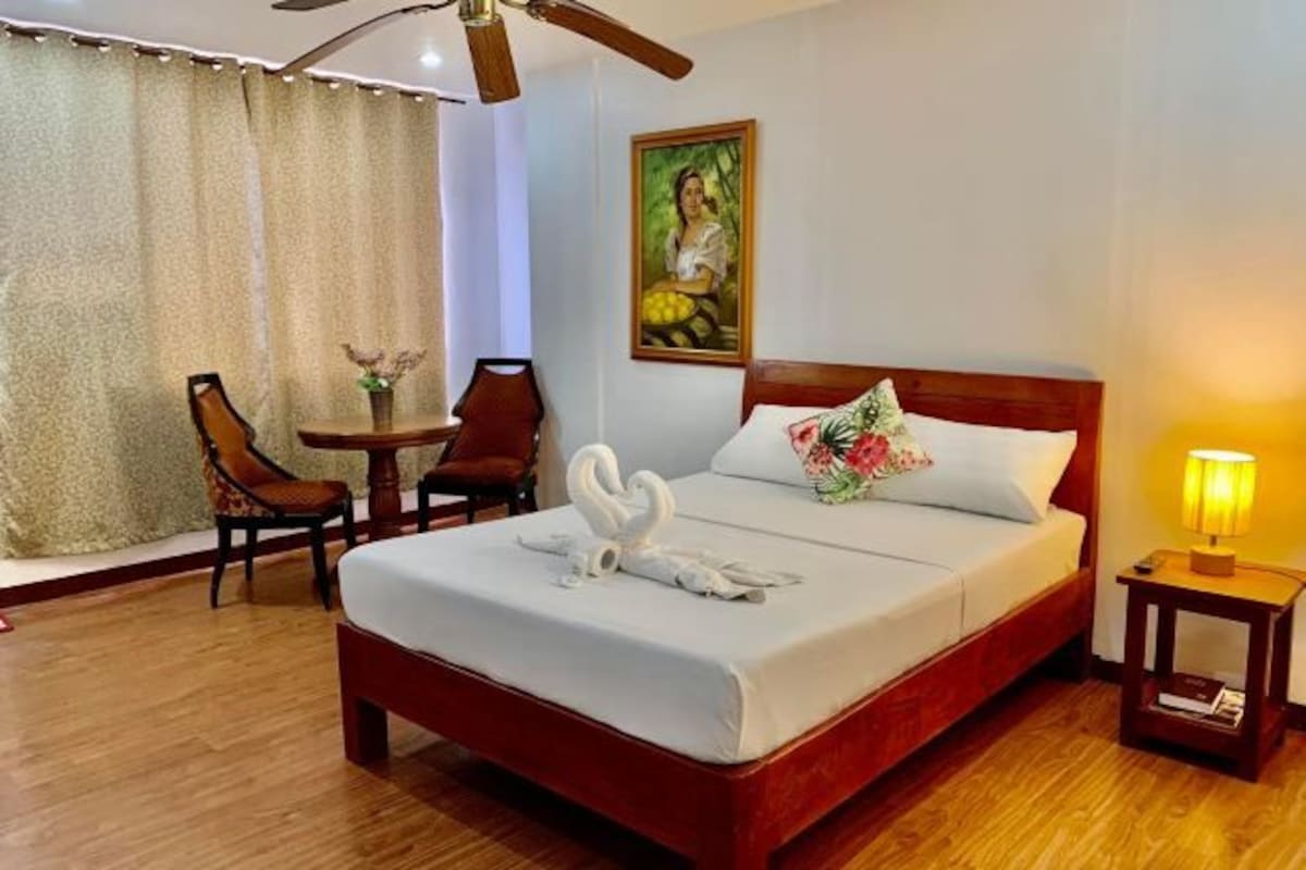 A hotel room with a wooden bed, a small table with chairs, a painting on the wall, and a ceiling fan.