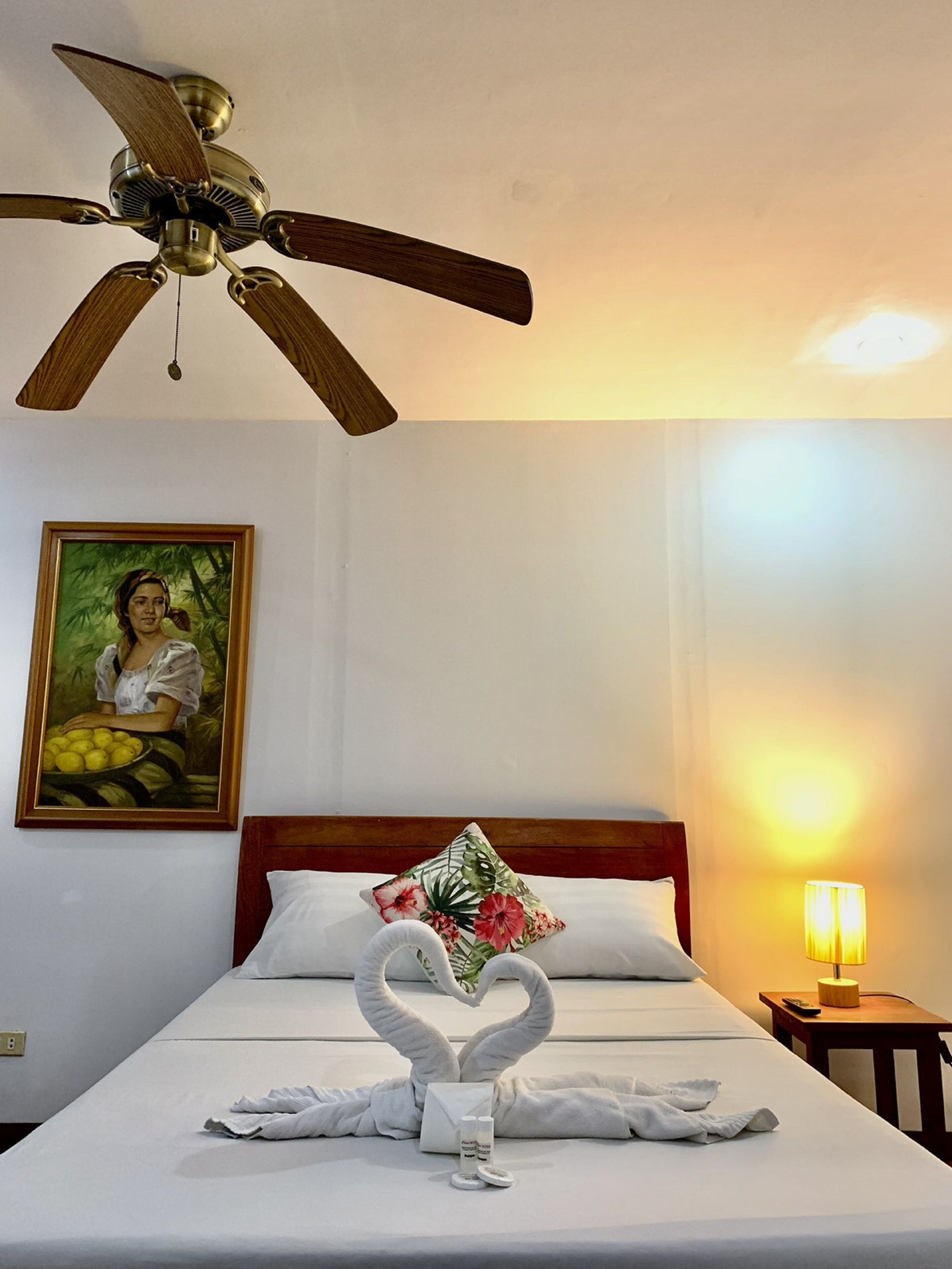 Habitación de hotel con cama, ventilador de techo, un cuadro, almohada con estampado floral, toallas dispuestas en forma de corazón y una lámpara de noche.