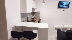 Eine kleine Küche mit weißer Arbeitsplatte, zwei schwarzen Barhocker, einem kleinen Esstisch und einem an der Wand befestigten Fernseher.