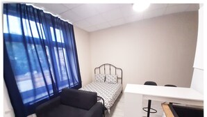 Ein kleiner, ordentlich eingerichteter Raum mit einem Bett, einem Schreibtisch, einem Sessel und einer Couch.