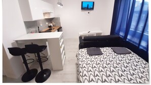 Ein modernes Zimmer mit Küchenzeile, einem Bett mit gemusterten Betttextilien, einem an der Wand montierten Fernseher und einem Fenster mit blauen Vorhängen.