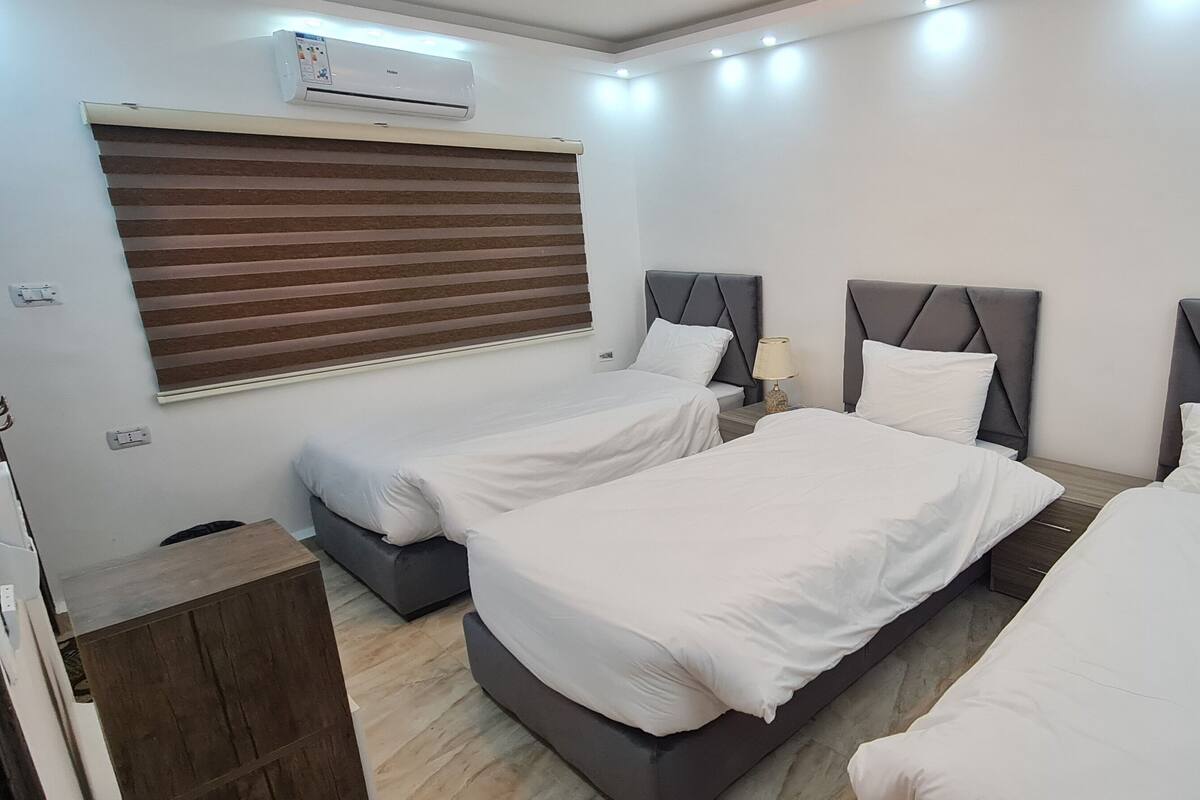 A hotel room with three beds, a window with blinds, an air conditioning unit, and a bedside table with a lamp.