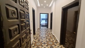 A hallway with patterned floor tiles, a door with decorative panels, and a view into a room with a bed and a cross on the wall. - Nabateans Home Hotel (Wadi Musa)