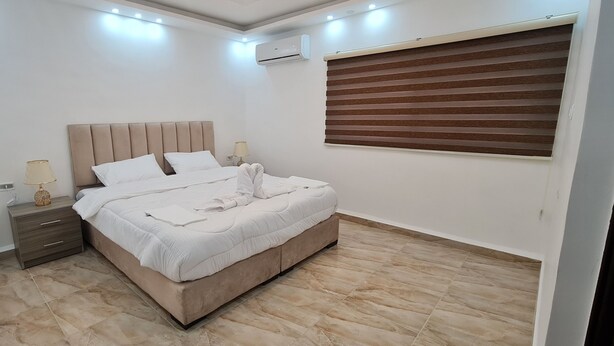 A bedroom with a bed, nightstands, a lamp, an air conditioner, and a window with blinds. - Nabateans Home Hotel (Wadi Musa)