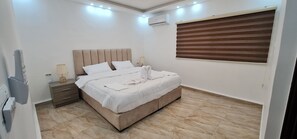 A bedroom with a bed, nightstands, a lamp, an air conditioner, and a window with blinds. - Nabateans Home Hotel (Wadi Musa)
