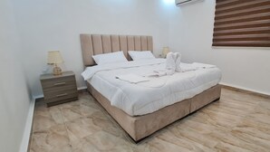 A neatly made bed with white linens and a beige headboard in a room with a tiled floor and a window with blinds. - Nabateans Home Hotel (Wadi Musa)