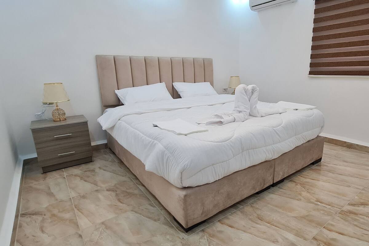 A neatly made bed with white linens and a beige headboard in a room with a tiled floor and a window with blinds.