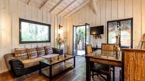 Ein Zimmer mit Holzboden, einer Couch mit gemusterten Kissen, einem Couchtisch, einem Esstisch mit Stühlen und einem an der Wand befestigten Fernseher.