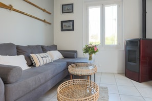Una sala de estar con un sofá gris, dos mesitas de mimbre, un jarrón con flores, una chimenea y cuadros en la pared.