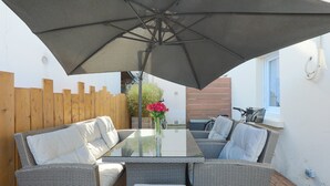Ein Tisch mit Glasplatte, Korbstühle, ein großer Sonnenschirm und ein Holzzaun auf einer Terrassenfläche.