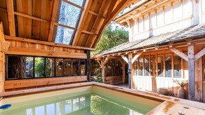A wooden spa with a large indoor pool, wooden beams, and a view of greenery outside.