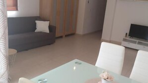 A modern living room with a glass dining table, white chairs, a grey sofa, and a TV stand.