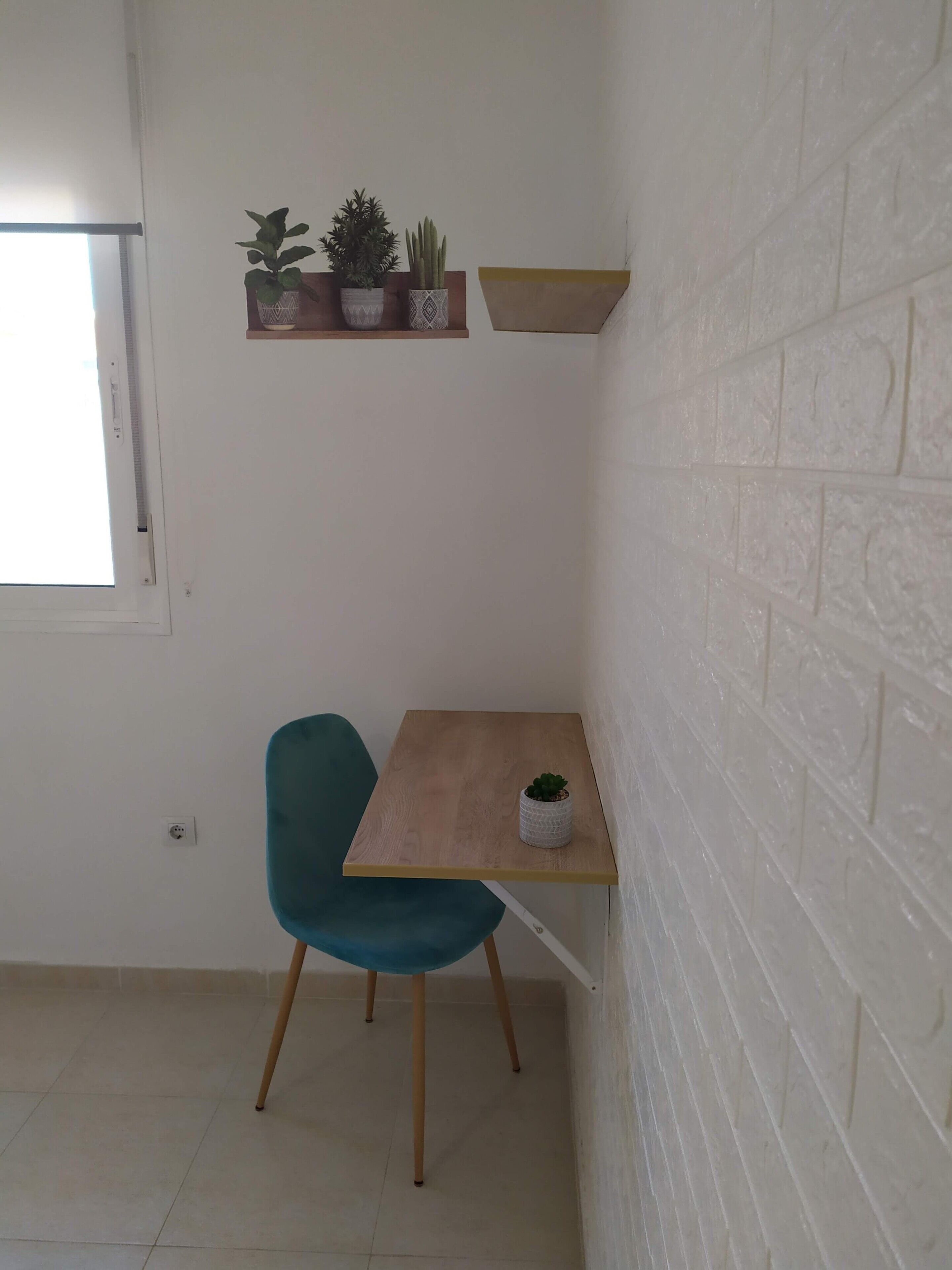 Una habitación pequeña con una mesa de madera, una silla verde azulada, una planta en maceta, una estantería con plantas y una pared de ladrillo blanco.