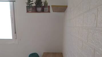 Una habitación pequeña con una mesa de madera, una silla verde azulada, una planta en maceta, una estantería con plantas y una pared de ladrillo blanco.