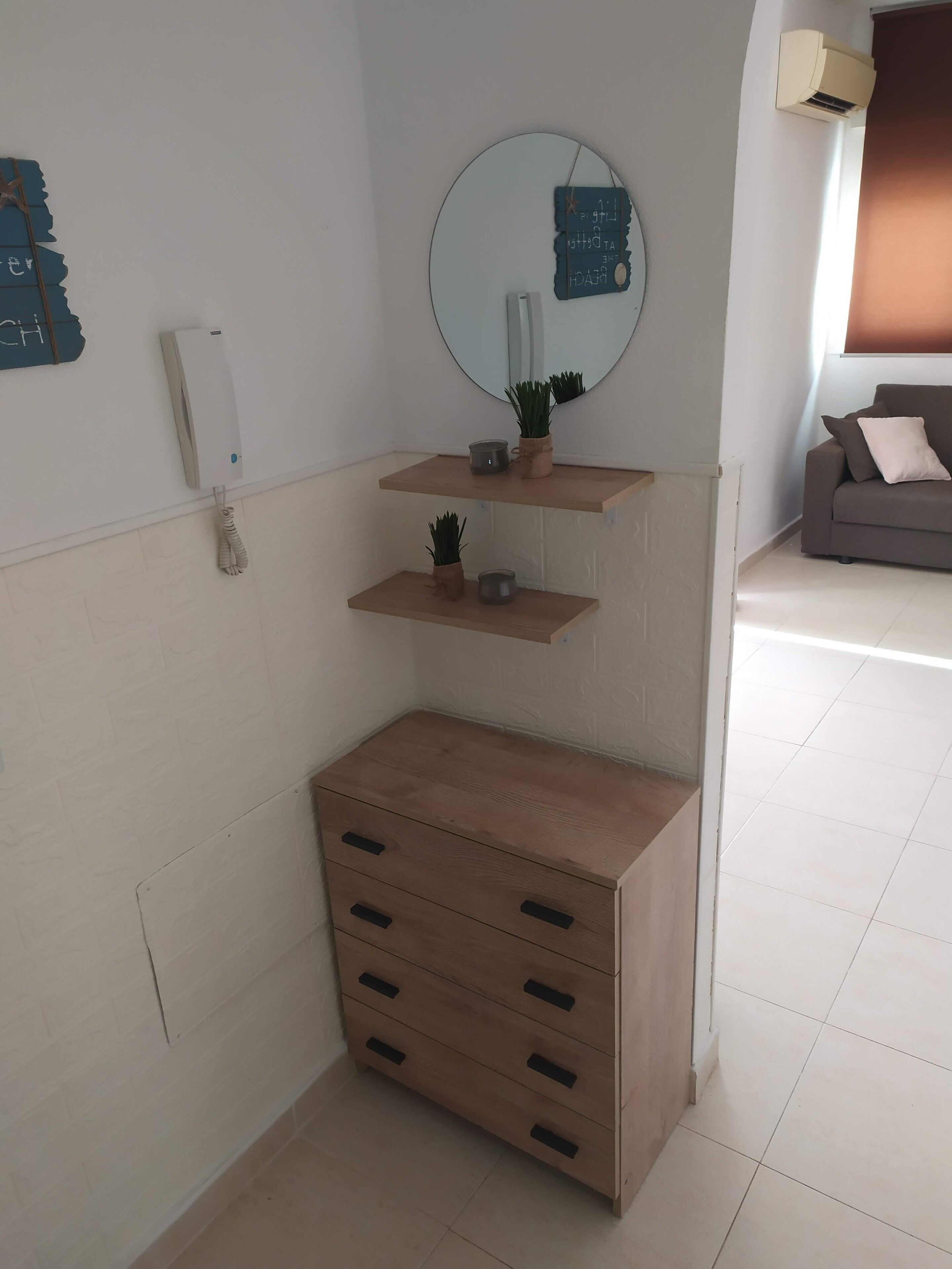 Une pièce d’angle avec un meuble en bois, un miroir fixé au mur et une petite étagère ornée d’objets décoratifs.