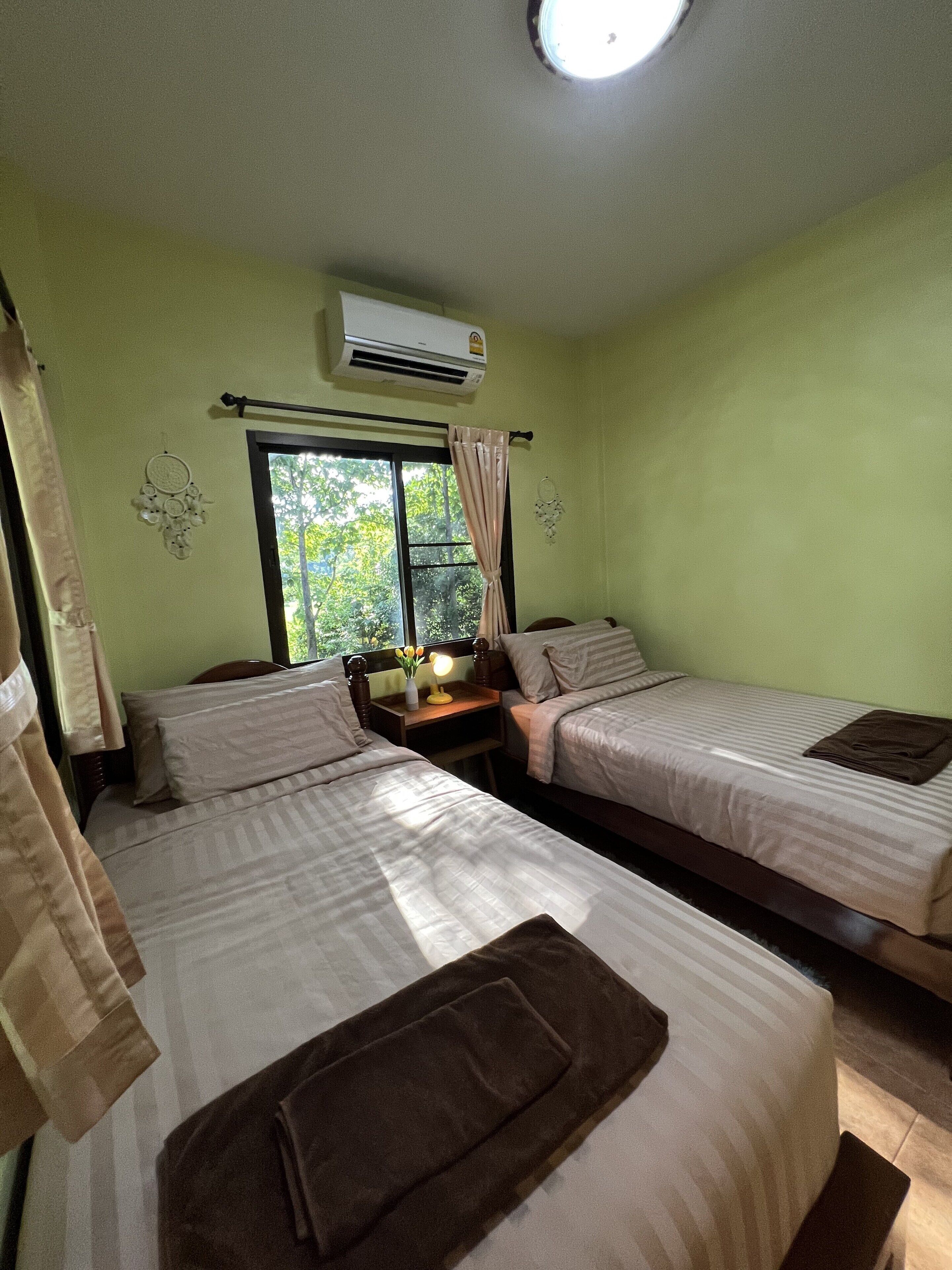 A room with two beds, a window with curtains, an air conditioning unit, and a bedside table with a lamp.