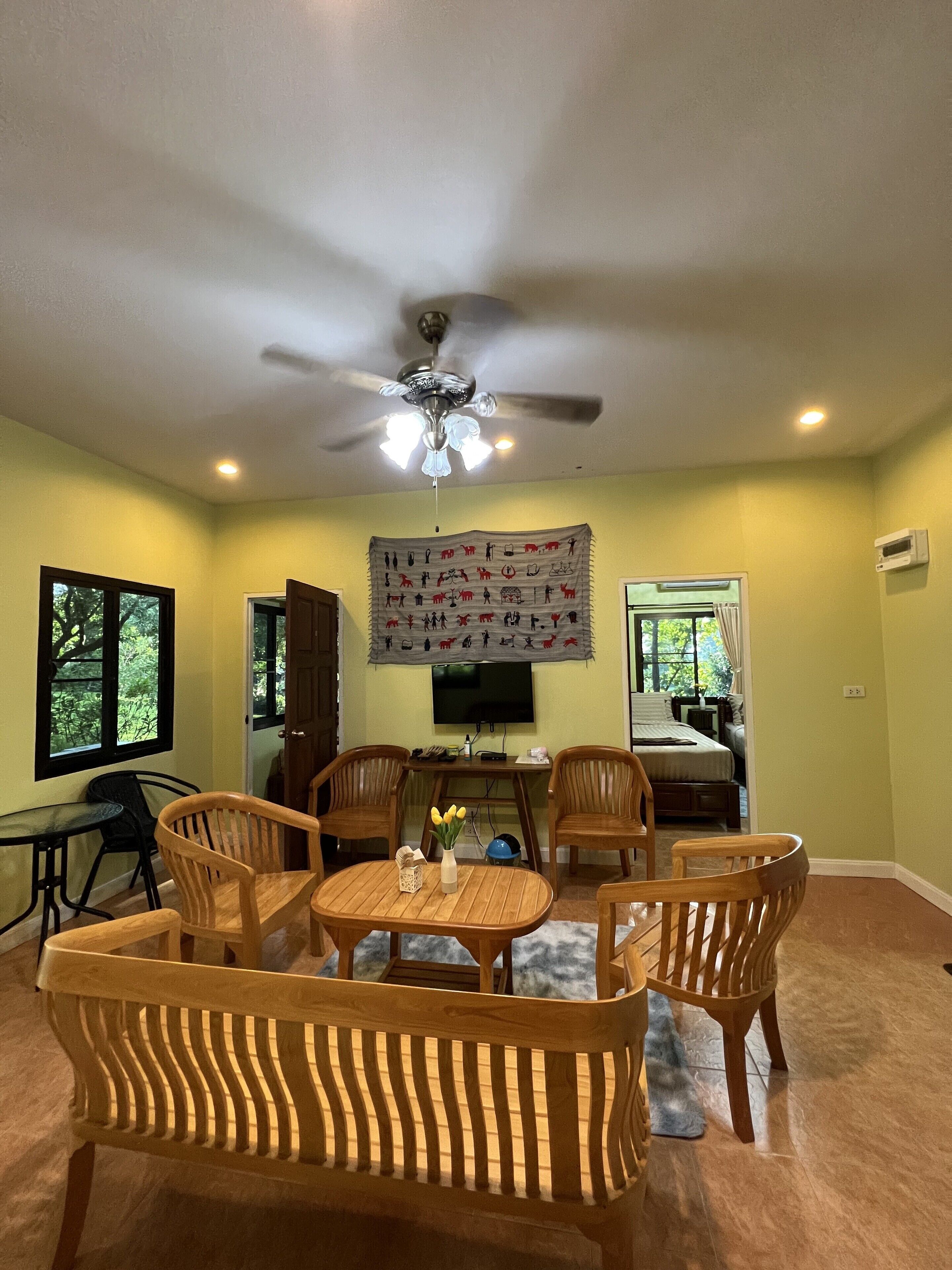 A living room with a ceiling fan, a dining table with chairs, a TV mounted on the wall, and a bed in the background.