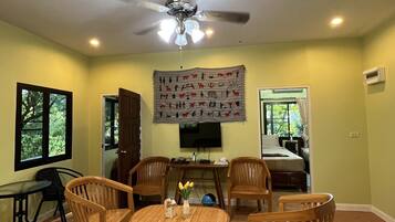 A living room with a ceiling fan, a dining table with chairs, a TV mounted on the wall, and a bed in the background.