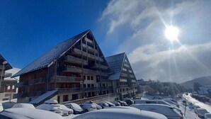 Ein mehrstöckiges Gebäude mit Balkonen, ein Parkplatz mit schneebedeckten Autos und ein klarer Himmel mit Sonnenschein.