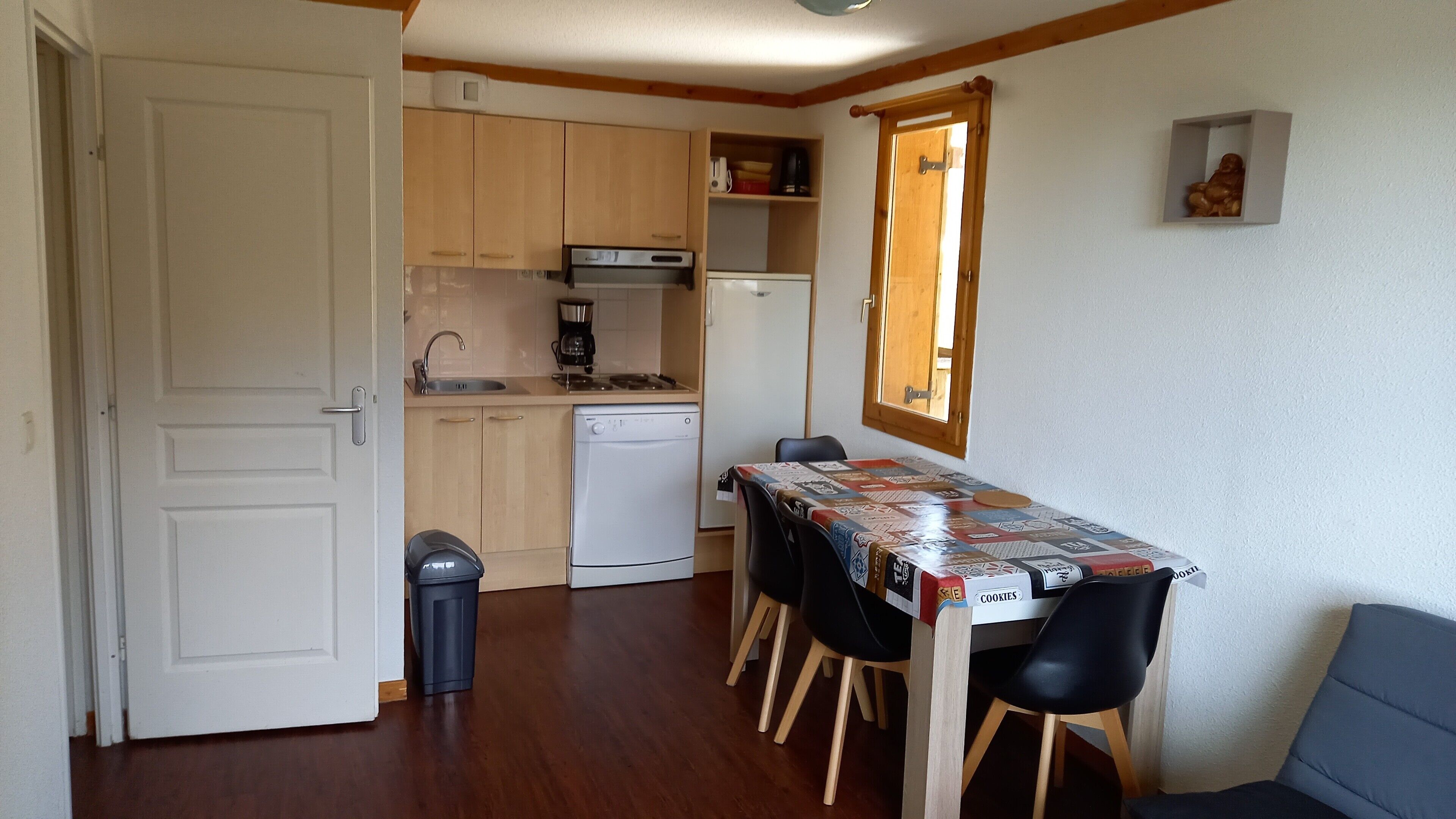 Een kleine keuken met houten kastjes, een vaatwasser en een eettafel met stoelen.