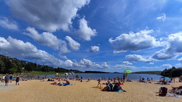 Ein Strand mit badenden und sonnenden Menschen, einem klaren Himmel und einem See im Hintergrund.