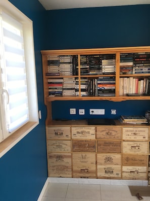 A room with a bookshelf filled with books and DVDs, a window with blinds, and wooden crates labeled with wine names.
