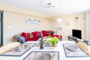 Un salon moderne avec une table à manger pour deux personnes, un canapé avec des coussins rouges, un téléviseur et une plante.