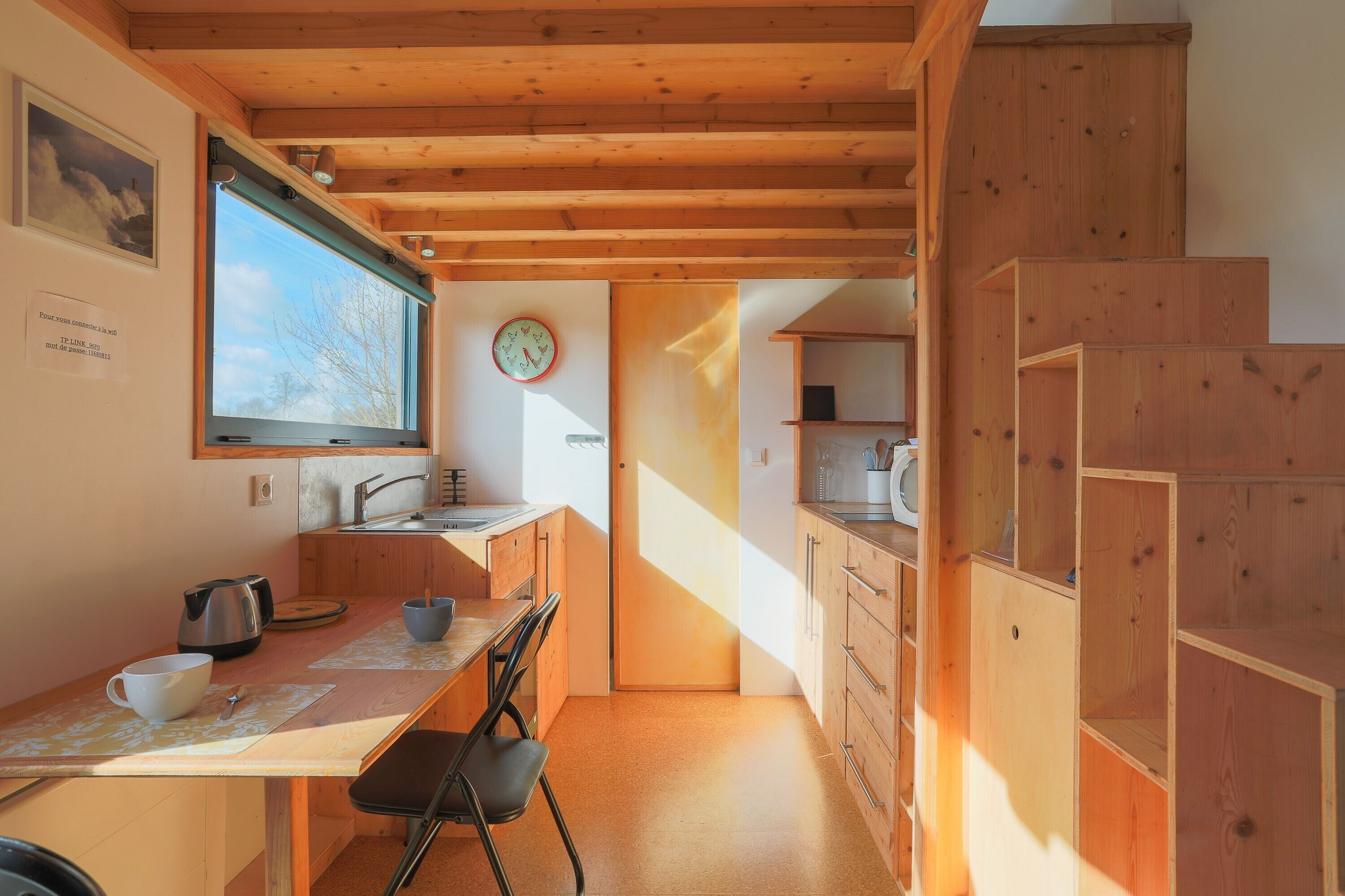 A kitchen with wooden cabinets, a dining table, chairs, a kettle, a clock, and a window.