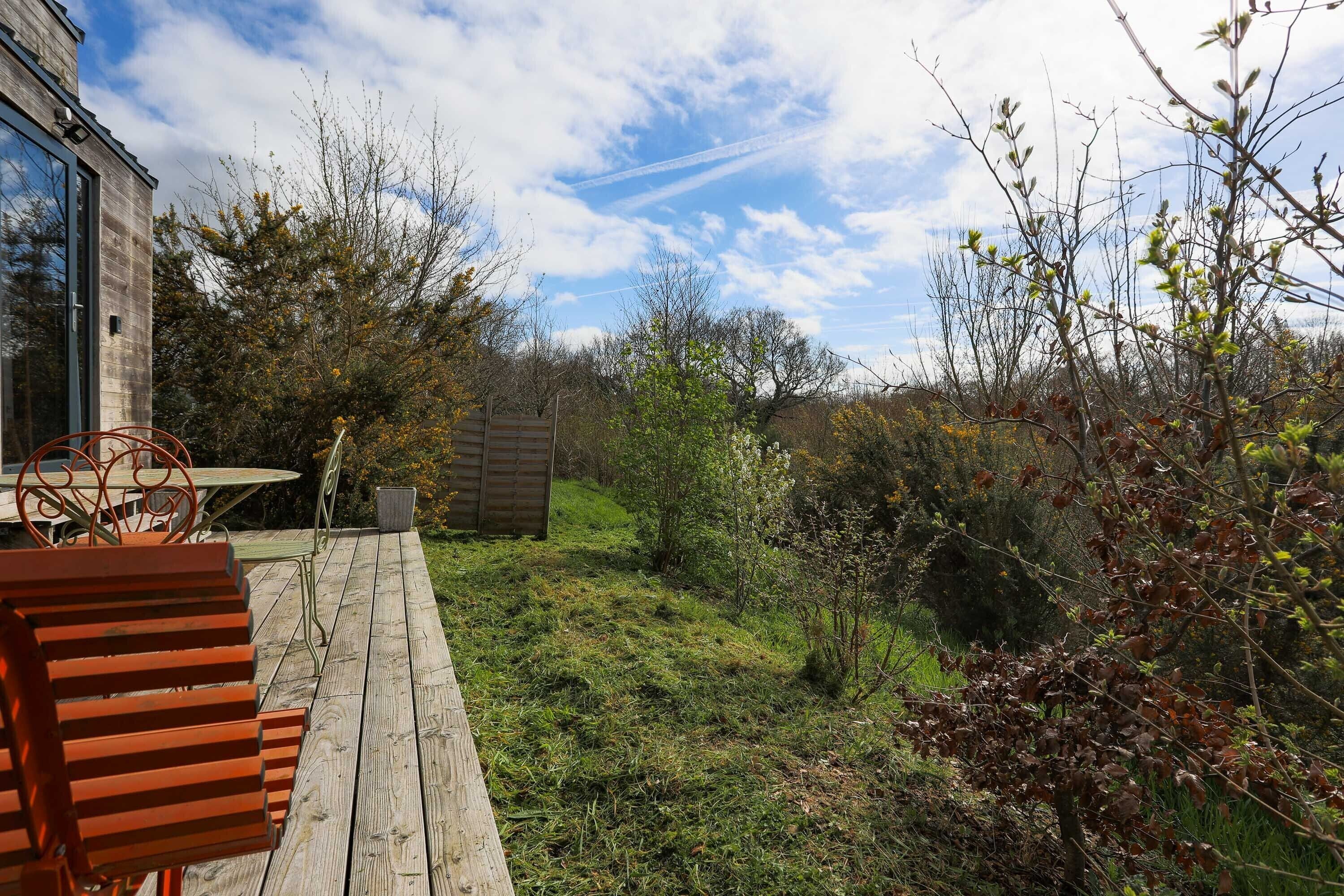 A wooden deck with a metal table and chairs, surrounded by greenery and trees.