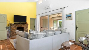 A living room with a stone fireplace, a grey sofa, a television, and a sliding glass door leading to an outdoor area. - Villa "Norgreen 4" with Private Pool, Private Terraces and Wi-Fi (Grimaud)