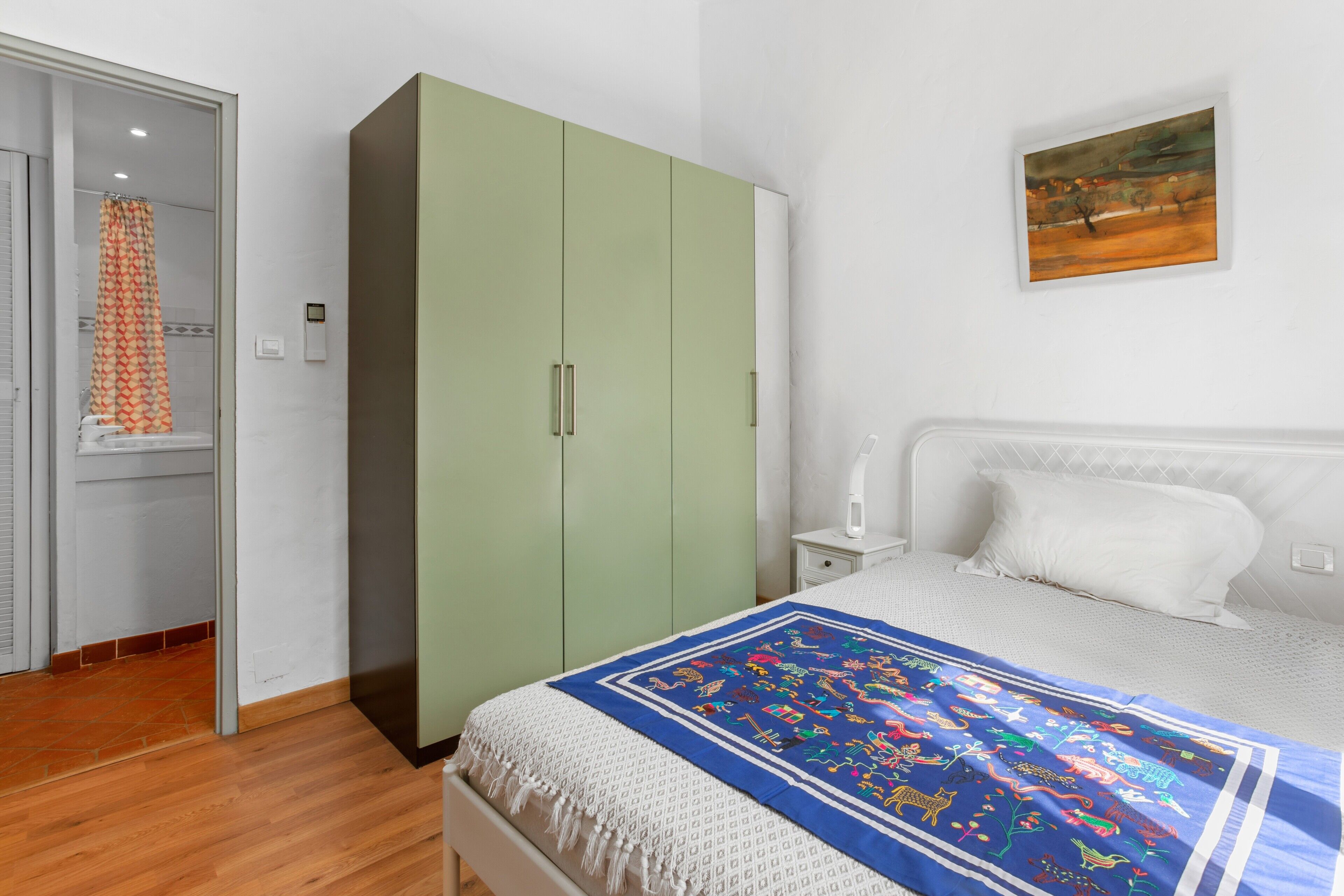 A bedroom with a bed, a wardrobe, a nightstand, a painting, and a bathroom with a shower curtain.