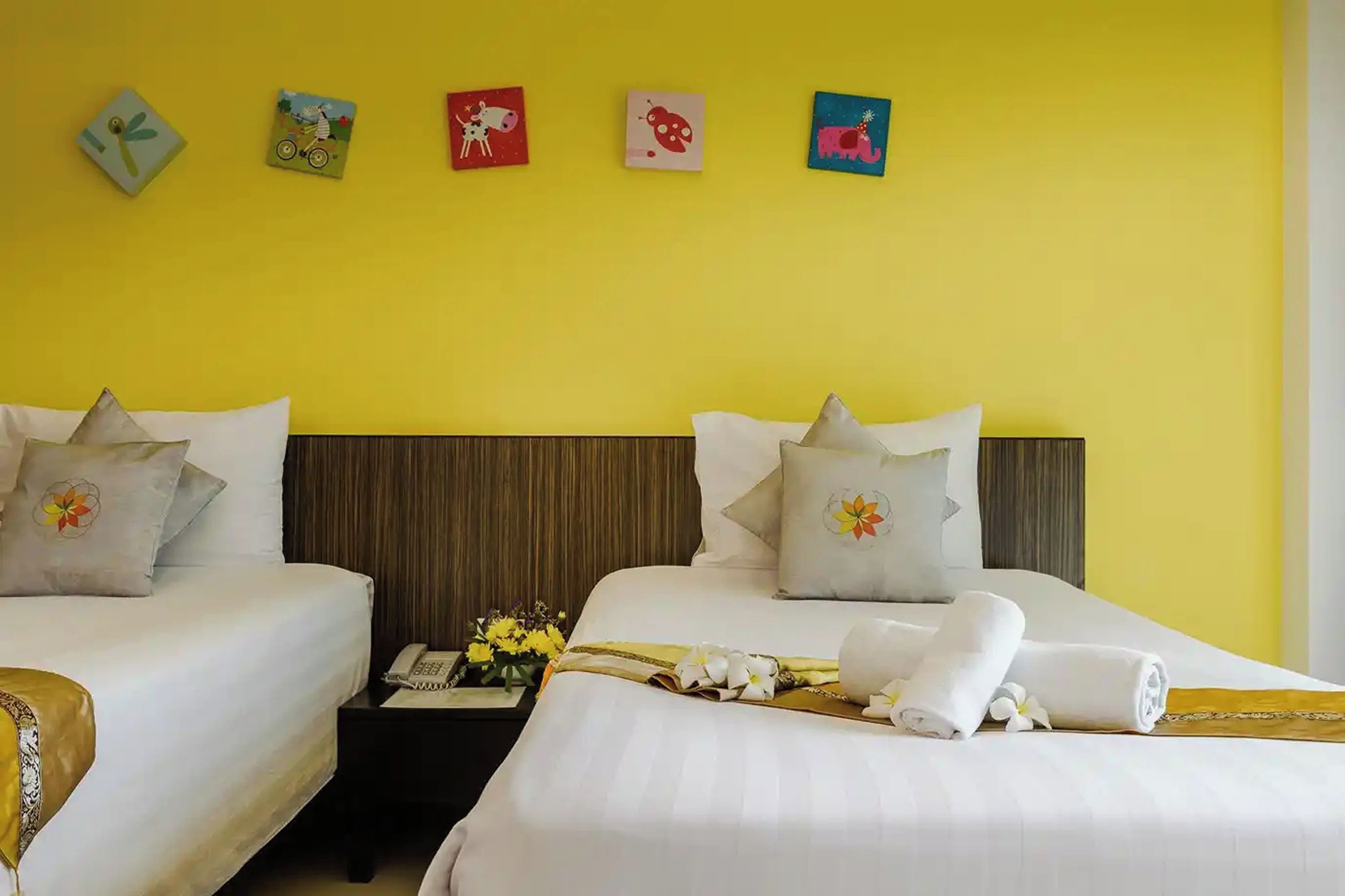 Habitación de hotel con dos camas, una mesita de noche con un jarrón de flores y una pared decorada con obras de arte de colores.