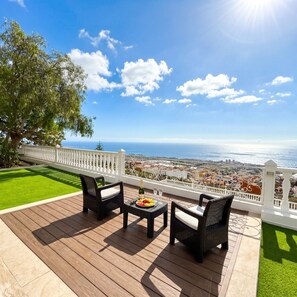 A terrace with wicker furniture, a table with a bottle and fruit, and a view of the sea and town.
