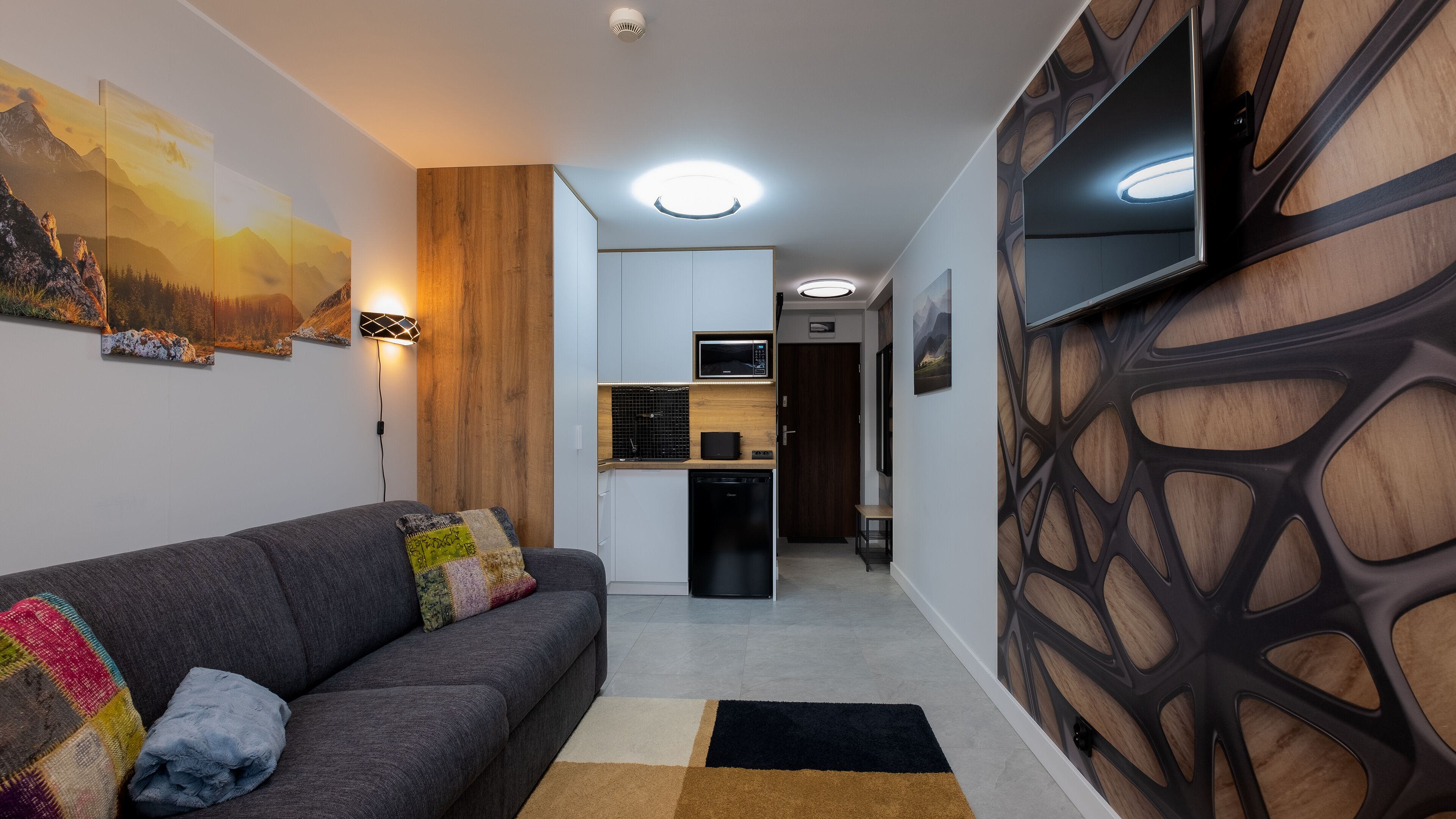 Una sala de estar moderna con un sofá gris, cojines coloridos, un panel de madera decorativo y un televisor de pantalla plana montado en la pared.