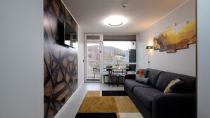 Una sala de estar moderna con un sofá gris, una mesa de centro y un televisor montado en la pared. Hay un balcón con vistas al exterior.