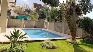 A rectangular swimming pool with a brick wall surrounding it, a grassy area with plants, and a building in the background. - Myra Residences by Extended Stays 2 Bedrooms (Nairobi)