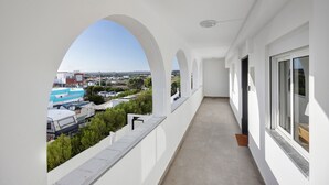Un pasillo con paredes blancas y aberturas en arco, que conduce a un balcón con vistas a vehículos estacionados y edificios.