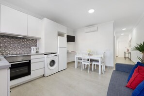 Une cuisine moderne dotée d’appareils électroménagers intégrés, un coin repas avec une table et des chaises, et un espace de vie avec un canapé et une plante.