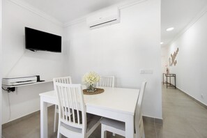 Un espace repas comprenant une table blanche, des chaises, un téléviseur à écran plat, un appareil de climatisation et une petite table ornée d’un vase de fleurs.