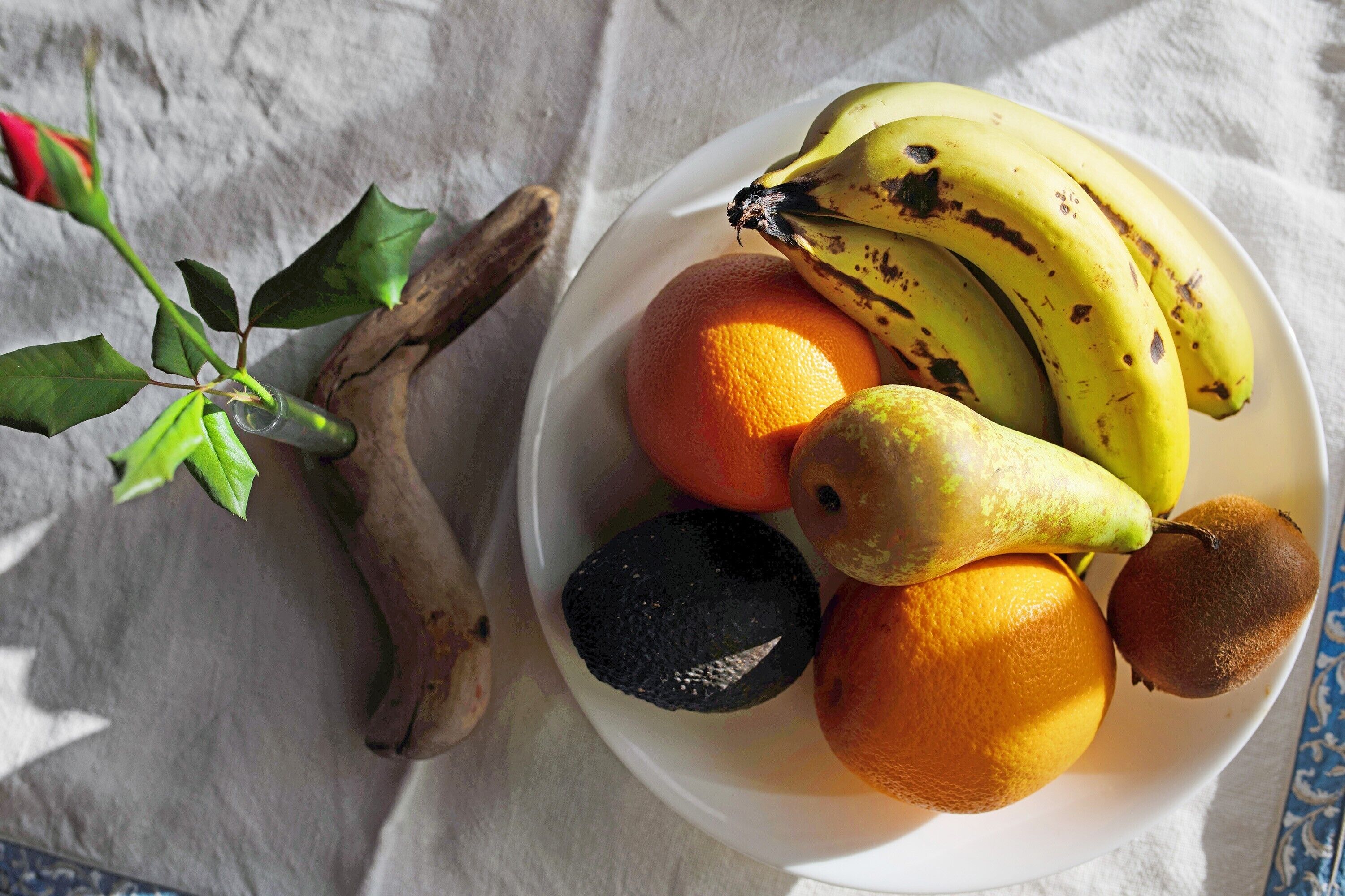 Un saladier rempli de fruits, notamment des bananes, des oranges et un kiwi, posé sur une table recouverte d’une nappe.