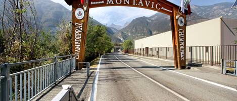 A wooden archway with 'Montavica' and 'Welcome' signs, a paved road, metal railings, and mountainous background.