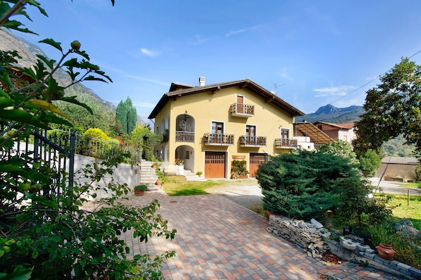Una casa de dos pisos con un balcón, un patio empedrado y montañas al fondo.