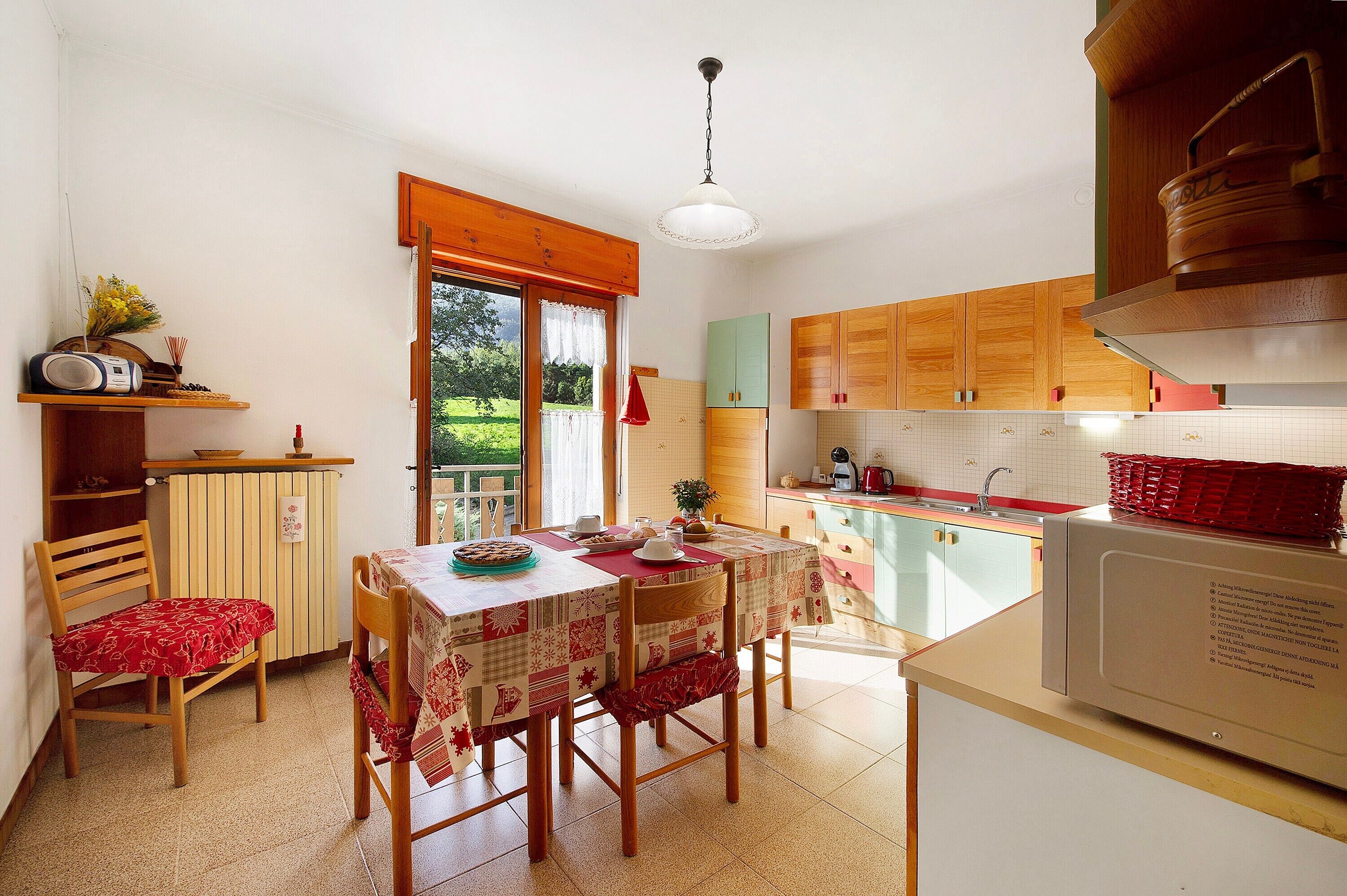 Een keuken met houten kastjes, een eettafel met stoelen, een magnetron en een raam met uitzicht op groen.