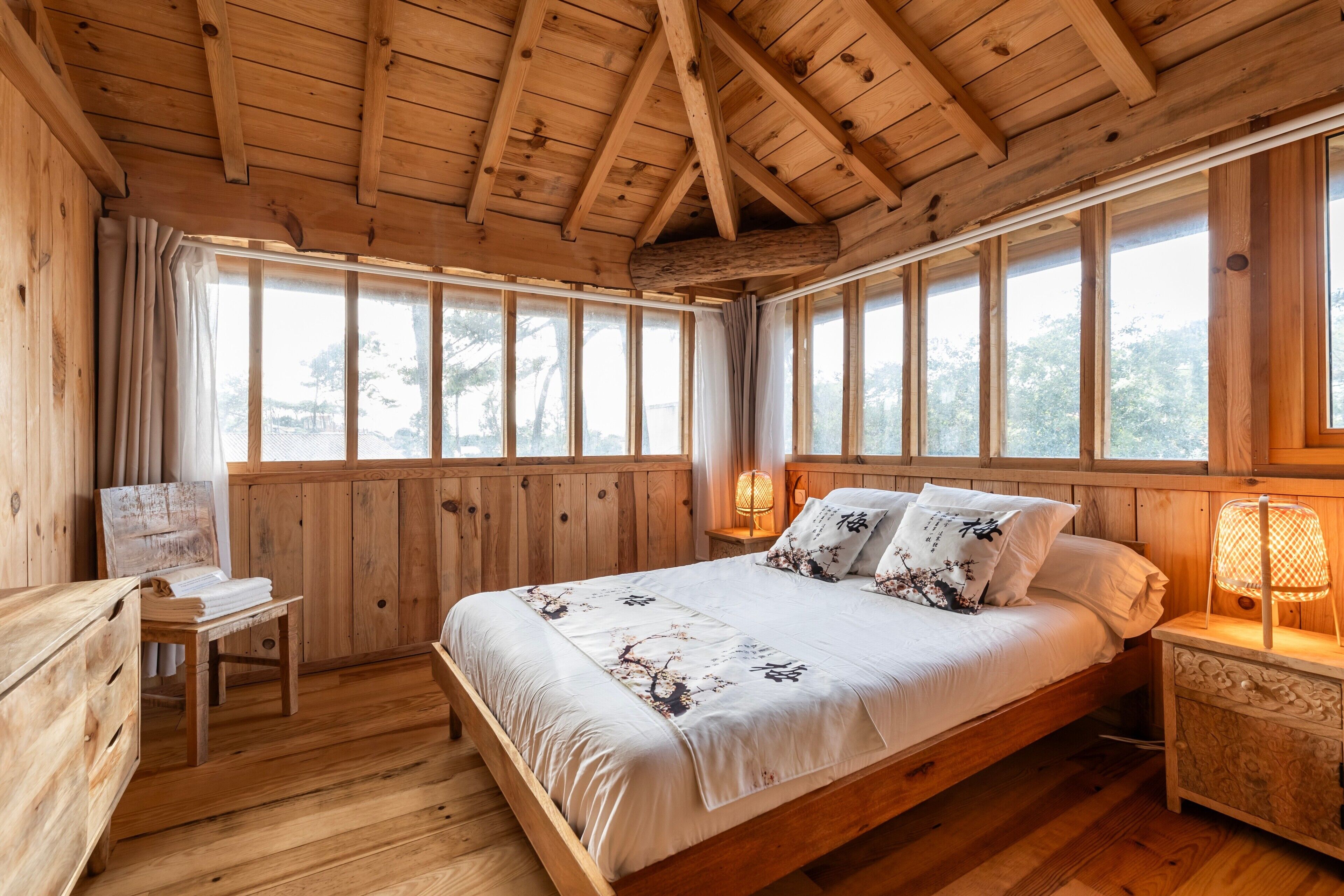 Une chambre dans une cabane en bois, avec un lit, des lampes de chevet, un bureau et de grandes fenêtres.